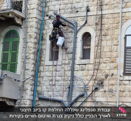 אדם תלוי על חבלים עם דלי וכלי עבודה ליד צינורות על קיר.